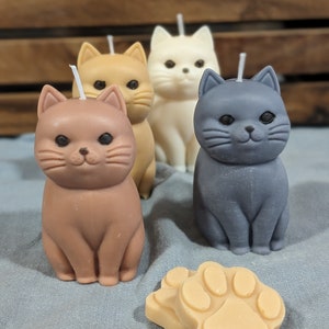 May include: Four cat-shaped candles, one brown, one orange, one white, and one gray, are arranged on a blue surface. A paw-shaped candle is in the foreground.