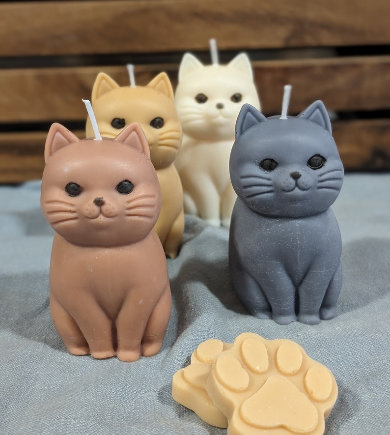 May include: Four cat-shaped candles, one brown, one orange, one white, and one gray, are arranged on a blue surface. A paw-shaped candle is in the foreground.