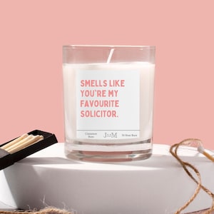 Puede incluir: Una vela blanca en un tarro de vidrio con una etiqueta rosa que dice "Smells like you're my favourite solicitor." La vela est&aacute; etiquetada como "Cinnamon Buns" y "30 Hour Burn".