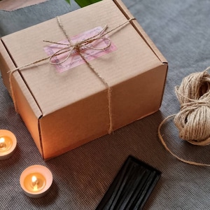 May include: A brown cardboard box tied with twine and a pink label that says "Candles". The box is sitting on a gray surface with two lit candles and a box of black matches.