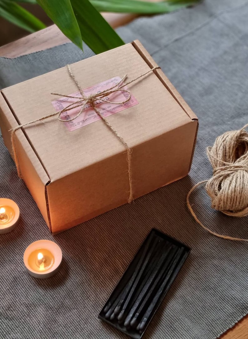 May include: A brown cardboard box tied with twine and a pink label that says "Candles". The box is sitting on a gray surface with two lit candles and a box of black matches.