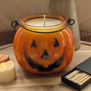 K&ouml;nnte beinhalten: Ein orangefarbener K&uuml;rbis-Kerzenhalter aus Glas mit schwarzem Gesicht und Metallgriff. Die Kerze im Inneren ist wei&szlig;. Eine Schachtel Streichh&ouml;lzer befindet sich neben dem Kerzenhalter. Der Kerzenhalter steht auf einer Holzoberfl&auml;che.