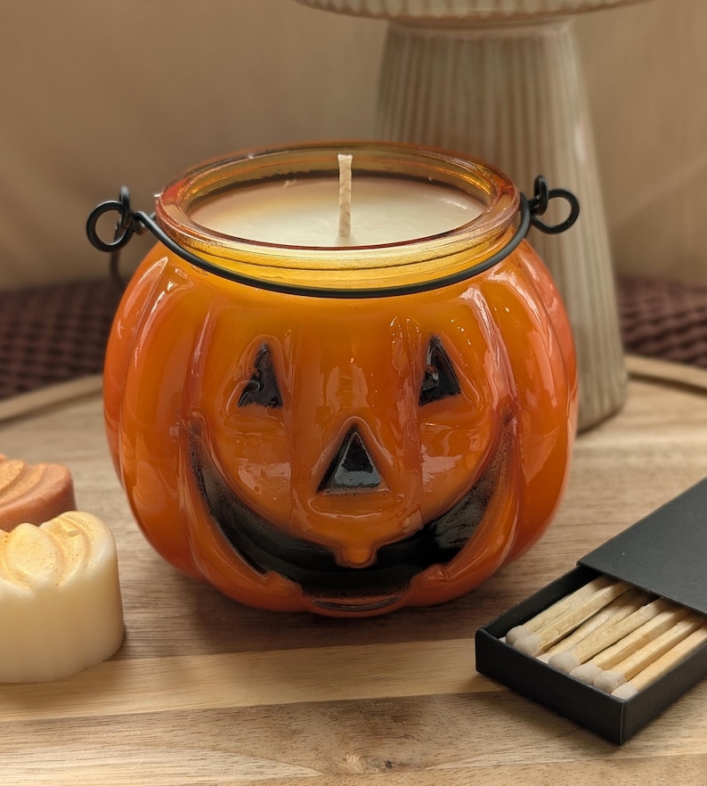 K&ouml;nnte beinhalten: Ein orangefarbener K&uuml;rbis-Kerzenhalter aus Glas mit schwarzem Gesicht und Metallgriff. Die Kerze im Inneren ist wei&szlig;. Eine Schachtel Streichh&ouml;lzer befindet sich neben dem Kerzenhalter. Der Kerzenhalter steht auf einer Holzoberfl&auml;che.