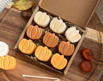 May include: A box filled with small pumpkin-shaped wax melts in various shades of orange and white. The box is open and sitting on a wooden surface with two matchsticks.