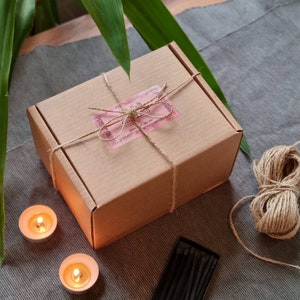 May include: A brown cardboard box tied with twine and a pink label that reads "Palm Candles". The box is sitting on a gray textured surface with two small candles and a box of matches.