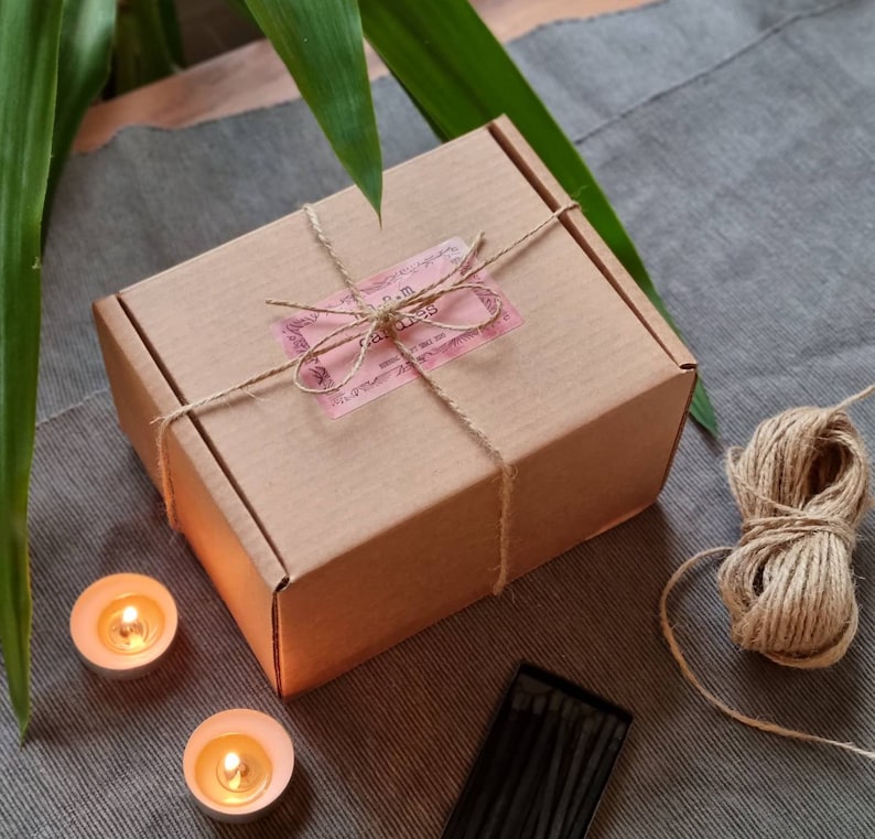 May include: A brown cardboard box tied with twine and a pink label that reads "Palm Candles". The box is sitting on a gray textured surface with two small candles and a box of matches.
