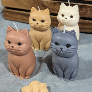 May include: Four cat-shaped candles in different colors: brown, beige, white, and gray. The candles are sitting on a blue fabric surface.