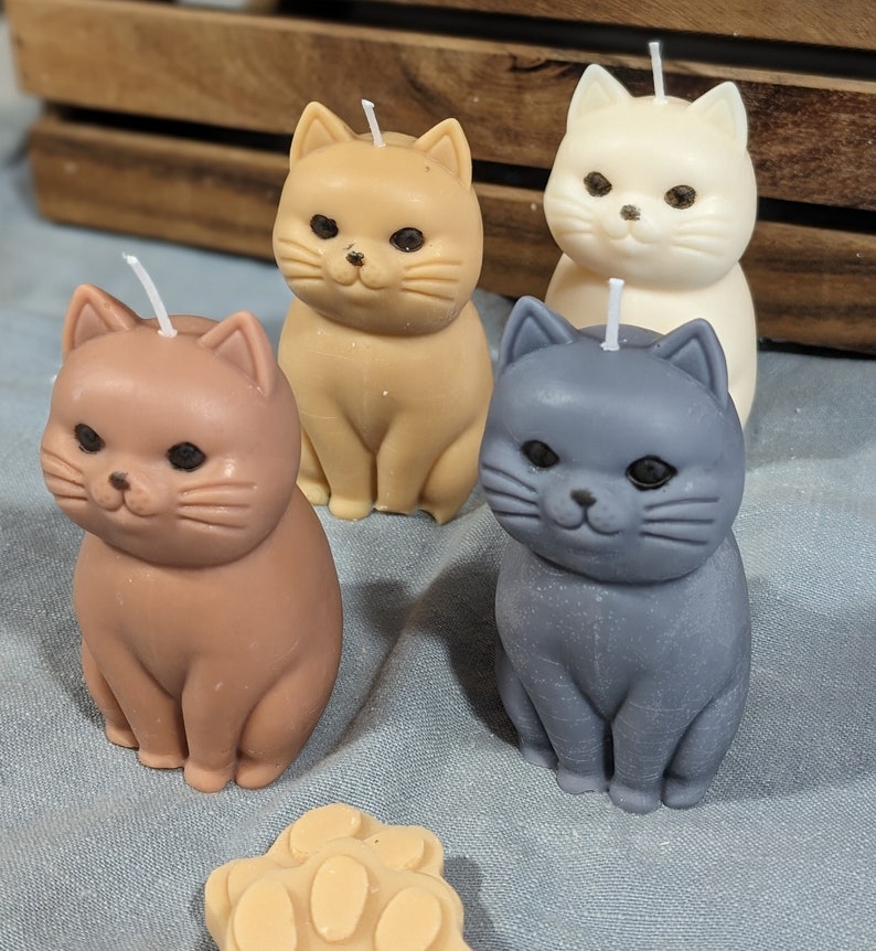 May include: Four cat-shaped candles in different colors: brown, beige, white, and gray. The candles are sitting on a blue fabric surface.