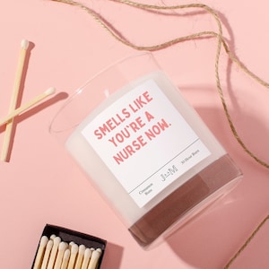 May include: A clear glass candle jar with a white label that reads "Smells like you're a nurse now." The label is pink and has a black border. The candle is cinnamon scented and has a 30-hour burn time. The candle is on a pink surface with a piece of twine and matches.