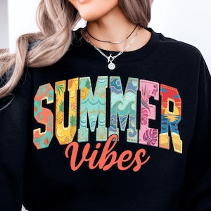 Puede incluir: Sudadera negra con la frase "SUMMER VIBES". La palabra "SUMMER" está en letras grandes con diseños tropicales. "Vibes" está en cursiva naranja. El diseño incorpora piñas, olas y puestas de sol.