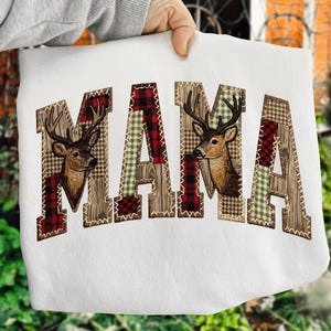 Op de afbeelding: Witte sweatshirt met het woord "MAMA" in een patchwork-ontwerp. De letters zijn versierd met ruitjes, houtnerf en groene gingham patronen. Twee hertenkoppen zijn geborduurd in de letters, wat een rustieke uitstraling geeft.