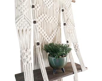 May include: A macrame wall hanging with a wooden shelf attached. The macrame is made with natural cotton cord and features a geometric pattern with wooden beads. A small potted plant sits on the shelf.