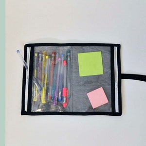 Op de afbeelding: Een zwarte ritszak met een transparant plastic venster, waarin verschillende pennen en twee plakbriefjes te zien zijn. De tas heeft een zwart handvat en de tekst "EASY-VIEW DOUBLE ZIPPER POUCH" staat bovenaan de afbeelding. De tekst "SAGE SEWING PATTERNS" staat onderaan de afbeelding.