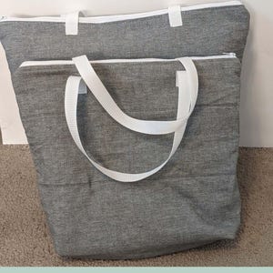 Pode incluir: Duas sacolas de compras cinza isoladas com alças e zíperes brancos. As sacolas têm o texto "INSULATED GROCERY BAG" na parte superior e "SAGE SEWING PATTERNS" na parte inferior. As sacolas são feitas de tecido acolchoado.