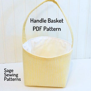 May include: A yellow and white striped fabric basket with a white lining and a yellow handle. The basket is empty and has a white background. The text "Handle Basket PDF Pattern" is written in black above the basket. The text "Sage Sewing Patterns" is written in black below the basket.