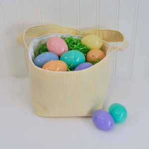 May include: A yellow striped fabric Easter basket filled with pastel plastic eggs. The basket has a white lining and a white handle. Two pastel plastic eggs are sitting outside the basket.