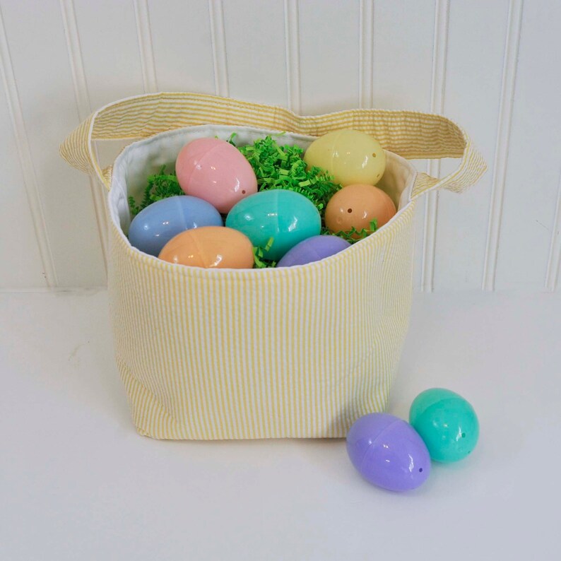 May include: A yellow striped fabric Easter basket filled with pastel plastic eggs. The basket has a white lining and a white handle. Two pastel plastic eggs are sitting outside the basket.