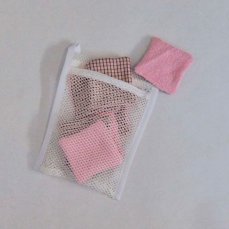 K&ouml;nnte beinhalten: Ein wei&szlig;er W&auml;schebeutel aus Netzstoff mit Rei&szlig;verschluss. Der Beutel enth&auml;lt pink- und schwarz gemustertes Stoff und einen rosa Waschlappen.