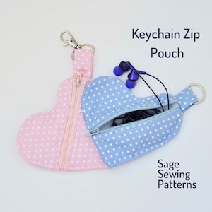 Może przedstawiać: Dwie serduszkokształtne saszetki zapinane na zamek błyskawiczny w kropki. Jedna saszetka jest różowa w białe kropki, a druga niebieska w białe kropki. Obie saszetki mają zamek błyskawiczny i kółko na klucze.