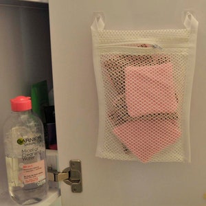 K&ouml;nnte beinhalten: Ein wei&szlig;er Netz-W&auml;schebeutel mit Rei&szlig;verschluss, der an einer wei&szlig;en Schrankt&uuml;r h&auml;ngt. Der Beutel enth&auml;lt rosa Abschminkpads. Eine durchsichtige Flasche Garnier SkinActive Micellar Cleansing Water ist auf der linken Seite des Bildes zu sehen.