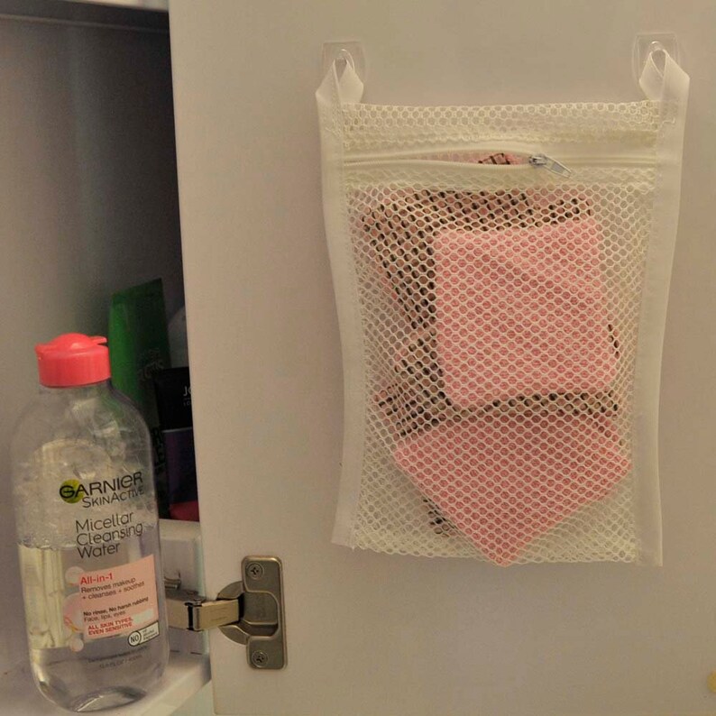 K&ouml;nnte beinhalten: Ein wei&szlig;er Netz-W&auml;schebeutel mit Rei&szlig;verschluss, der an einer wei&szlig;en Schrankt&uuml;r h&auml;ngt. Der Beutel enth&auml;lt rosa Abschminkpads. Eine durchsichtige Flasche Garnier SkinActive Micellar Cleansing Water ist auf der linken Seite des Bildes zu sehen.