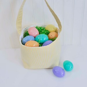 May include: A yellow and white striped fabric Easter basket filled with pastel plastic eggs. The basket has a fabric handle and is sitting on a white surface.