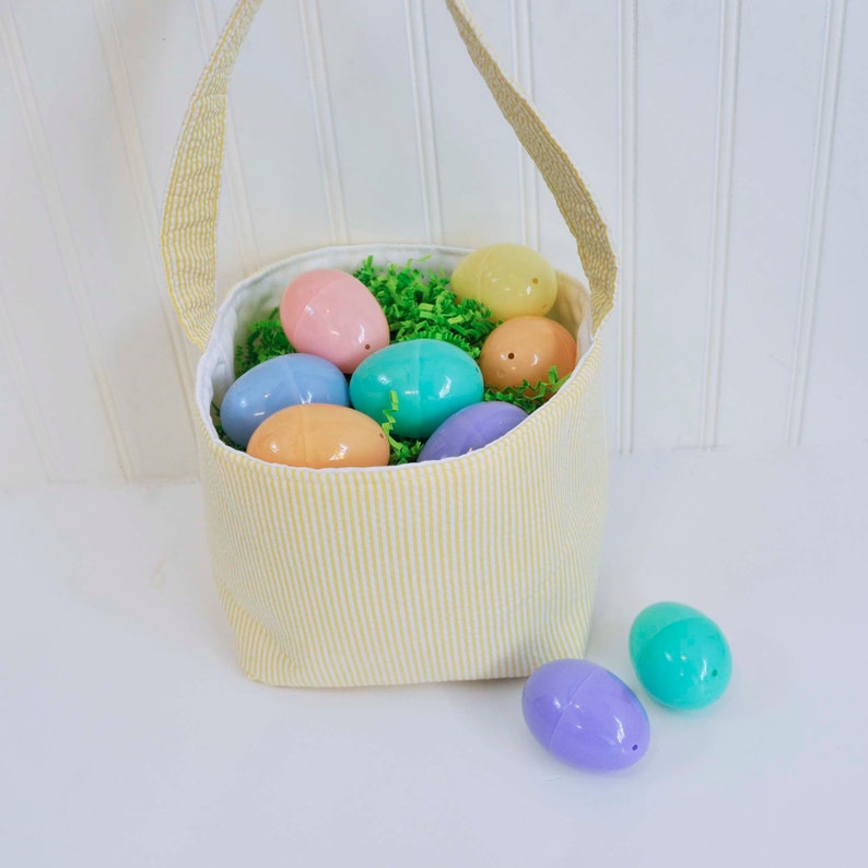 May include: A yellow and white striped fabric Easter basket filled with pastel plastic eggs. The basket has a fabric handle and is sitting on a white surface.