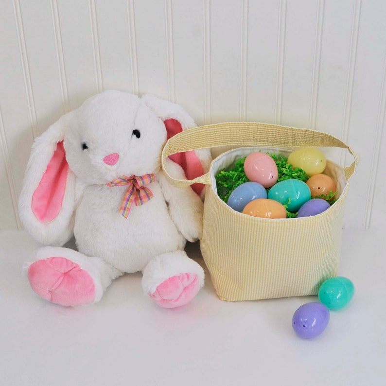 May include: A white plush bunny rabbit with pink ears sits next to a yellow striped fabric Easter basket filled with pastel colored plastic eggs.