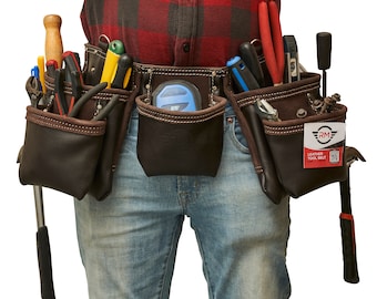 May include: A brown leather tool belt with multiple pockets and various tools, including a hammer, pliers, screwdrivers, and a tape measure. The belt is worn by a person wearing a red and black plaid shirt and blue jeans. The belt has a label that reads "RM Leather Tool Belt".