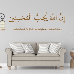 Op de afbeelding: Een beige bank met twee kussens, een decoratieve vogelkooilamp en een houten bijzettafel met een vaas met witte bloemen. De muur achter de bank heeft een bruine islamitische kalligrafie-quote in het Arabisch die luidt: "Voorwaar, Allah houdt van de weldoeners." De Engelse vertaling van de quote staat onder de Arabische tekst.