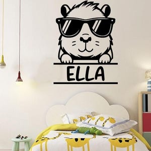 Könnte beinhalten: Schwarz-weißer Wandaufkleber mit einem Cartoon-Tier mit Sonnenbrille. Das Tier hält ein Banner mit dem Namen "ELLA". Entworfen für ein Kinderzimmer.