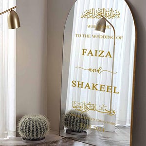 Puede incluir: Un espejo dorado con la parte superior arqueada, que presenta un anuncio de boda para Faiza y Shakeel. El anuncio incluye caligrafía árabe y el texto "Bienvenido a la boda de Faiza y Shakeel" y "Bendecidos en parejas 22 2024".