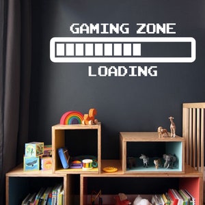 May include: A white vinyl wall decal with the text "GAMING ZONE" and a progress bar that reads "LOADING". The decal is on a dark gray wall above a set of colorful wooden shelves with toys and books.
