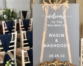 May include: A gray frosted acrylic sign with a gold decorative frame. The sign reads "Welcome to the Walimah of Wasim & Mashooda 28.08.22".
