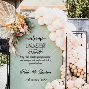 May include: A welcome sign with a green background and black text. The sign features an Arabic inscription, a message wishing the couple well, and the names "Ruba & Linkon" with the date "26th October 2022". The sign is decorated with white balloons.