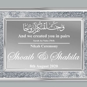 May include: A silver framed mirror with a glittery border. The mirror has a white inscription that reads "And we created you in pairs" in English and Arabic. The inscription also includes the verse "Surah An Naba (78:8)" and the words "Nikah Ceremony". The names "Shoaib & Shakila" and the date "8th August 2020" are also inscribed on the mirror.