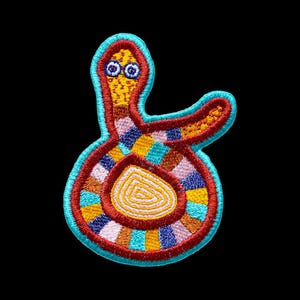 May include: An embroidered patch featuring a stylised creature with a yellow head, two blue eyes, and a coiled body. The design incorporates a vibrant patchwork pattern of red, orange, yellow, light blue, and dark blue segments with a bright teal border.