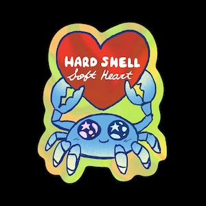 May include: A holographic sticker featuring a blue crab with star-shaped pupils holding a large red heart. The text HARD SHELL Soft Heart is printed in white across the heart. The sticker has a wavy, iridescent border against a black background.