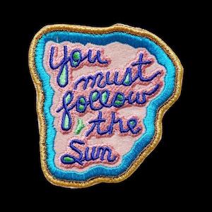 May include: An embroidered patch with a light pink center featuring the dark blue cursive text You must follow the Sun. The design includes small green accents, a bright blue border, and a metallic gold outer edge on a black background.