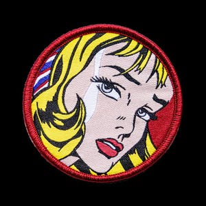 Op de afbeelding: Een ronde geborduurde patch met een pop-art illustratie van een persoon met blond haar, rode lippen en een traan op de wang. Het ontwerp is omrand door een dikke rode gestikte rand tegen een effen zwarte achtergrond.