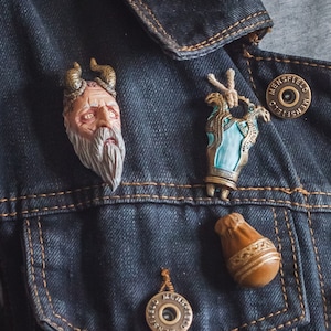 Puede incluir: Tres pequeños alfileres esculpidos detallados en una chaqueta vaquera azul. Los alfileres son una cara esculpida con cuernos, una mano esculpida sosteniendo una botella y una bolsa esculpida.