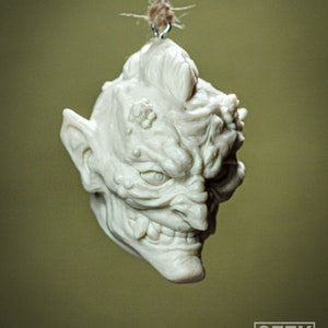 May include: A white ceramic ornament shaped like a goblin head with a mischievous expression. The ornament is hanging from a string.