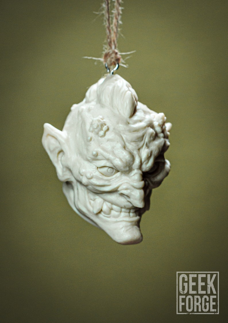 May include: A white ceramic ornament shaped like a goblin head with a mischievous expression. The ornament is hanging from a string.