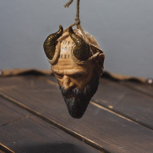 May include: A small, brown, ceramic sculpture of a man's head with horns and a beard. The head is covered in runes and is hanging from a string.