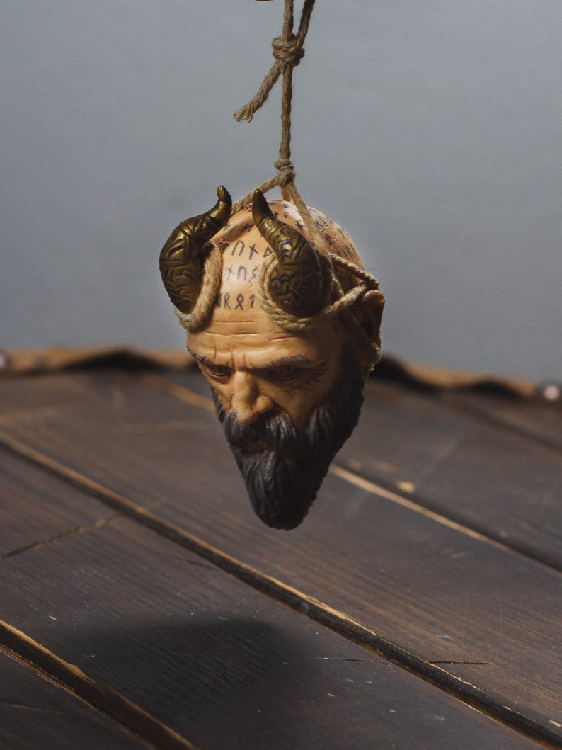 May include: A small, brown, ceramic sculpture of a man's head with horns and a beard. The head is covered in runes and is hanging from a string.