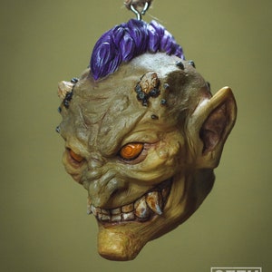 May include: A green and purple goblin head ornament with orange eyes and sharp teeth. The ornament is hanging from a string.