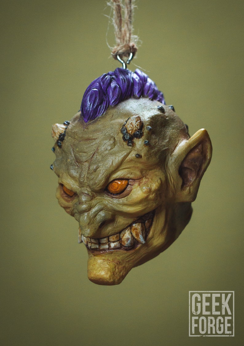 May include: A green and purple goblin head ornament with orange eyes and sharp teeth. The ornament is hanging from a string.