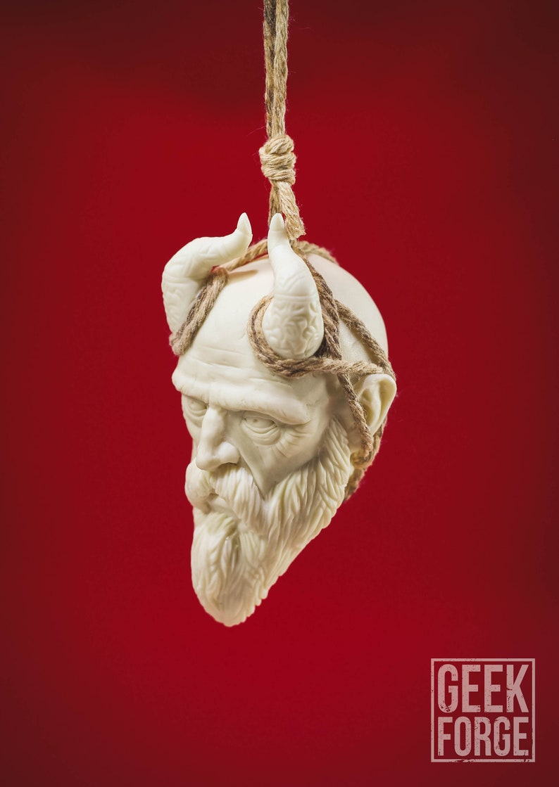 May include: A white ceramic sculpture of a man's head with horns and a beard, tied with twine and hanging from a rope against a red background.