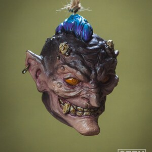 May include: A small, detailed, hand-painted ornament of a grotesque, goblin-like creature with a blue mohawk, orange eyes, and a gold nose ring. The ornament is hanging from a string.