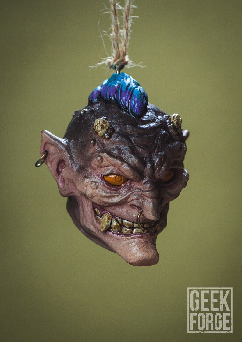 May include: A small, detailed, hand-painted ornament of a grotesque, goblin-like creature with a blue mohawk, orange eyes, and a gold nose ring. The ornament is hanging from a string.